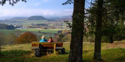 Two people sitting on a bench looking at the Vulkaneifel landskape